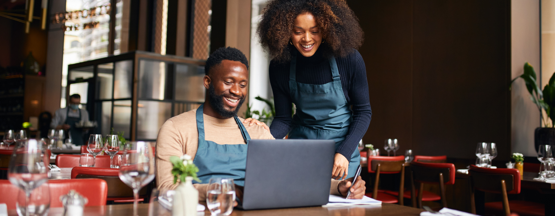 A restaurant manager in an apron is seated at a table in a restaurant looking at a laptop, while another worker looks over his shoulder.